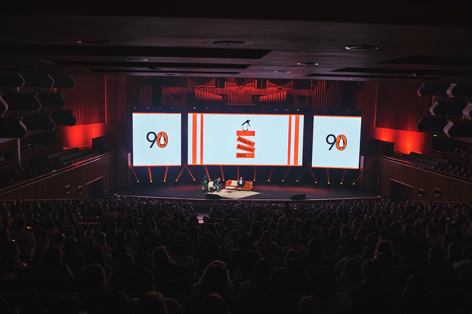 A packed auditorium facing the main stage at Penguin Presents, flanked by 90th anniversary screens